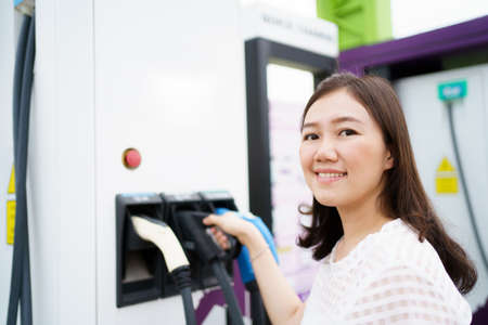 Happy Asian woman holding a DC - CCS type 2 EV charging connector at EV charging station, woman preparing an EV - electric vehicle charging connector for recharge a vehicle.の写真素材