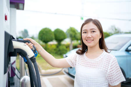 Happy Asian woman holding a DC - CCS type 2 EV charging connector at EV charging station, woman preparing an EV - electric vehicle charging connector for recharge a vehicle.の写真素材