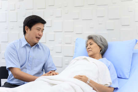 Senior Asian female patient resting on the medical bed in hospital and talking to her son. Son visiting and talking to his mother who staying in recovery ward.の写真素材