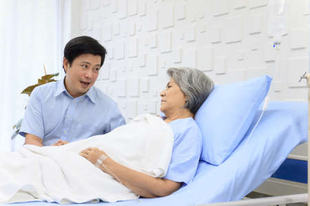 Senior Asian female patient resting on the medical bed in hospital and talking to her son. Son visiting and talking to his mother who staying in recovery ward.の写真素材