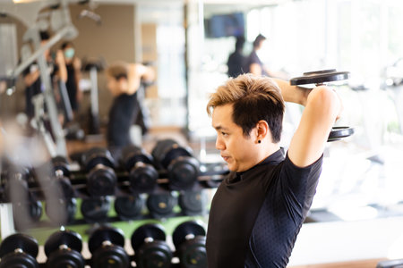 Active Asian sportsman doing a weight exercise in a gym - fitness by lifting up a dumbbell, man lifting up or holding a heavy dumbbell in Overhead Tricep Extension position. Bodybuilding workout.の写真素材