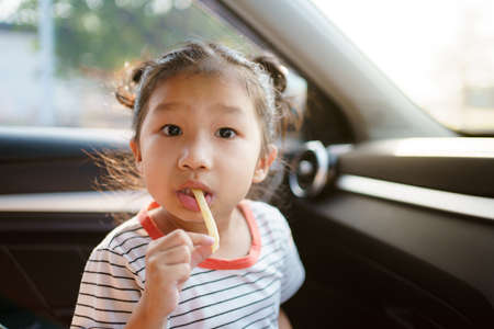 Little Asian young girl staying in the car and eating fried potato chips, lovely girl enjoy eating snack while traveling by the car.の写真素材