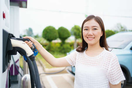 Happy Asian woman holding a DC - CCS type 2 EV charging connector at EV charging station, woman preparing an EV - electric vehicle charging connector for recharge a vehicle.の写真素材