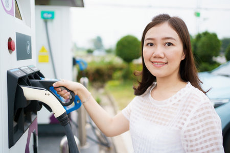 Happy Asian woman holding a DC - CCS type 2 EV charging connector at EV charging station, woman preparing an EV - electric vehicle charging connector for recharge a vehicle.の写真素材