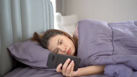 Lazy Asian young woman laying down on the bed and texting in social network with a friends close up.の写真素材