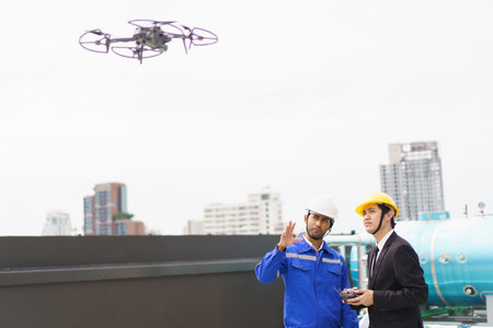 Professional Asian civil engineers using the unmanned aerial vehicle or UAV - drone for inspecting the building structure, 2 Asian foreman flying a drone to the building roof top area.の写真素材