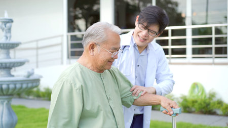 Happy senior Asian elder man trying to walk on the grass field to practice in physical therapy with a doctor. Very old man walking on the grass field with barefoot and using staff.の写真素材