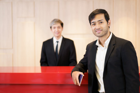 Senior Asian manager or chief executive officer - CEO standing at the reception desk in hotel with reception staff in background. Businessman in business traveling concept.の写真素材