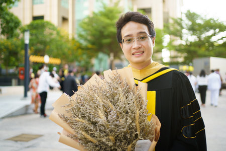Happy Asian graduate student holding a beautiful bouquet of flowers in the university graduation ceremony. Master degree student in gown suit showing a bouquet of flower for photography.の写真素材