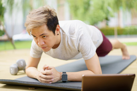 Active sportsman doing a plank exercise and making a video call with a friend and online sport training coach online. Healthy sport man making a body building workout with online instructor.の写真素材