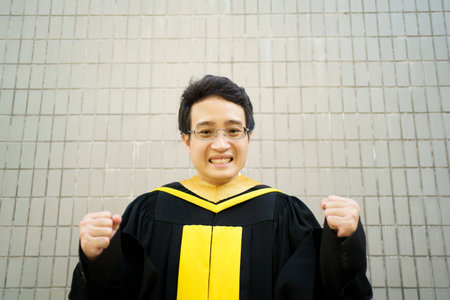 Happy Asian man in master degree gown in graduate ceremony. Male graduate student in gown holding showing a gladful face.の写真素材