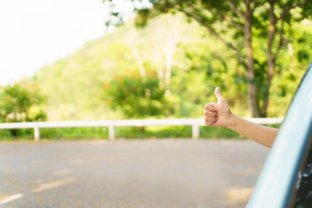 Unrecognizable Asian woman driver showing a thumb up outside the vehicle while driving a car. Happy woman driver or passenger showing her thumb up during in the car. Safety trip and freedom concept.の写真素材