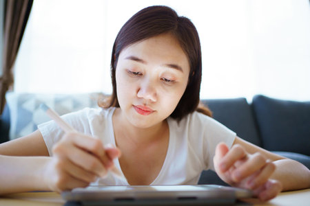 Happy Asian young woman relaxing in the living room and watching a contents and chatting with her friends and drawing an illustration on tablet. Woman enjoy watching video and drawing on tablet.の写真素材
