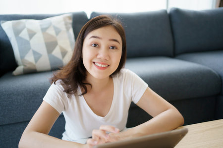 Happy Asian young woman relaxing in the living room and watching a contents and chatting with her friends and drawing an illustration on tablet. Woman enjoy watching video and drawing on tablet.の写真素材