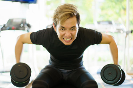 Active Asian sportsman doing a weight exercise in a gym - fitness by lifting up a dumbbell, man lifting up or holding a heavy dumbbell from dumbbell rack.の写真素材