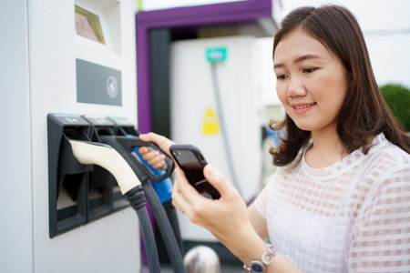 Happy Asian woman holding a DC - CCS type 2 EV charging connector at EV charging station, woman preparing an EV - electric vehicle charging connector for recharge a vehicle.の写真素材