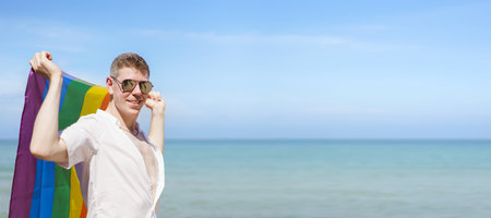Happy caucasian white man holding the rainbow flag or LGBT rainbow color flag while at the beach. Handsome man standing at the beach and showing LGBT flag.の写真素材