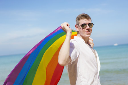 Happy caucasian white man holding the rainbow flag or LGBT rainbow color flag while at the beach. Handsome man standing at the beach and showing LGBT flag.の写真素材