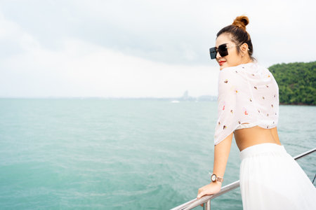 Happy beautiful and sexy Asian young woman in bikini posing for a photograph on the yacht deck. Sexy Asian lady in bikini standing on yacht deck and enjoy watching a beautiful view of sea and islands.の写真素材