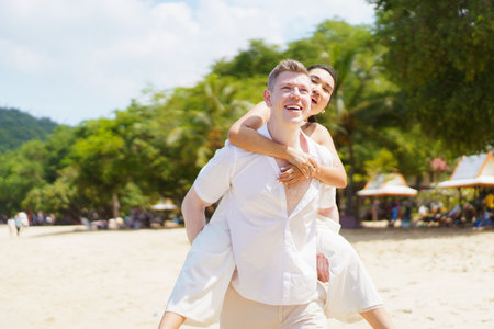 Happy Asian and caucasian white couple enjoy relaxing together at the beach during vacation or their honeymoon dating. Man piggyback or carrying his girlfriend on back and running on the beach.の写真素材