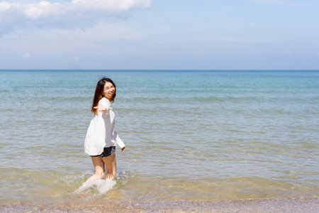 Happy Asian young woman enjoy playing and splashing a sea water at the beach. Summertime holiday and vacation concept. Cheerful Asian woman walking along the beach in morning and looking at camera.の写真素材