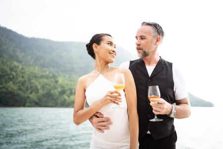 Happy Asian and caucasian white couple celebrating their marriage anniversary together on the yacht. Adult couple sailing a yacht and drinking a Champaign together during a honeymoon trip.の写真素材