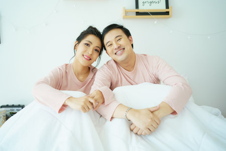 Happy and romantic Asian couple's portrait in bedroom with natural light from window, concept of relationship between husband and wife and being a family.の写真素材