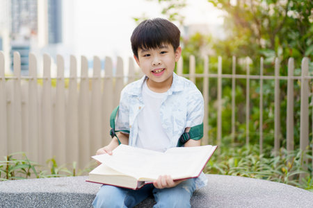 Cheerful happy Asian little boy sitting on the bench and reading a book or fairy tale at the public park alone.の写真素材