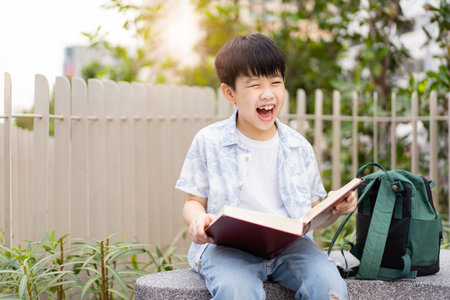 Cheerful happy Asian little boy sitting on the bench and reading a book or fairy tale at the public park alone.の写真素材