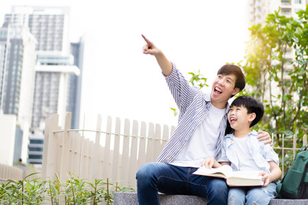 Cheerful happy Asian little boy and his father reading a book or fairy tale together at the outdoor park. Asian man and his son reading a book together.の写真素材