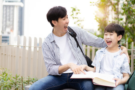 Cheerful happy Asian little boy and his father reading a book or fairy tale together at the outdoor park. Asian man and his son reading a book together.の写真素材