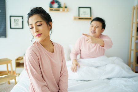 Unhappy - stressful Asian man sitting on the bed while his wife shouting at him. Critical or serious arguing between husband and wife about a problem in family.の写真素材