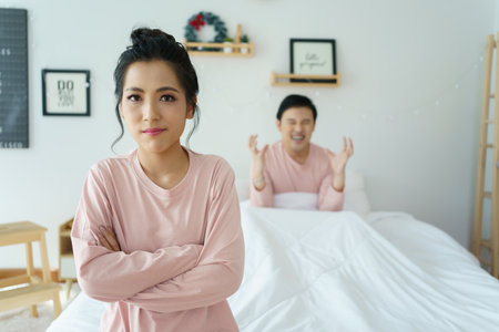 Unhappy - stressful Asian woman sitting and crying on the bed while her husband sleeping, a family and post marriage problem. Arguing Asian young couple with bad - negative relationship.の写真素材