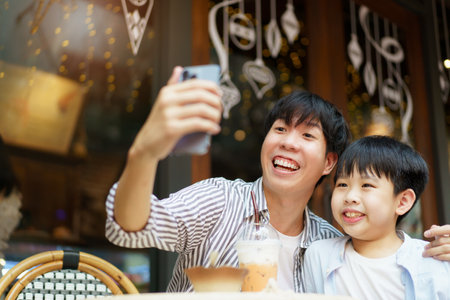 Happy Asian father and son enjoy a selfie photography or video call with their family and friends together while traveling. Cheerful young boy taking and taking a photo on smartphone with his dad.の写真素材