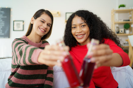 Caucasian white and African black ethnic women having a drinking together in celebrating event in their apartment bedroom. Woman colleague cheering and clinking a cocktail bottles.の写真素材