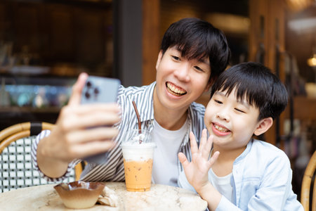 Happy Asian father and son enjoy a selfie photography or video call with their family and friends together while traveling. Cheerful young boy taking and taking a photo on smartphone with his dad.の写真素材
