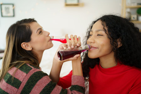 Caucasian white and African black ethnic women having a drinking together in celebrating event in their apartment bedroom. Woman colleague cheering and clinking a cocktail bottles.の写真素材