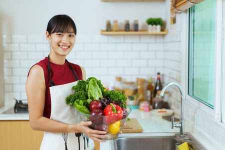 Happy healthy Asian woman washing a vegetables in domestic kitchen faucet before cooking. Cheerful healthy Asian woman carries a basket of vegetables and fruit and smiles to camera.の写真素材