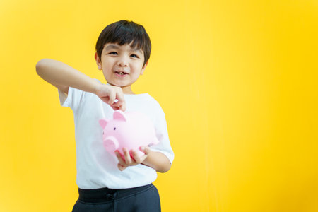 Happy Asian little young boy inserting or putting a coin into a saving piggy on a bright yellow background. Asian boy smiling and trying to put a coin into a piggy saving.の写真素材