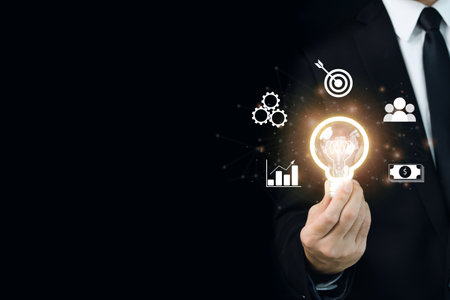 Unrecognizable good posing businessman holding a lightbulb in a hand showing the illuminated lightbulb and icons and network illustrations. Business development and invention concept.の写真素材