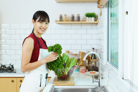 Happy healthy Asian woman washing a vegetables in domestic kitchen faucet before cooking. Cheerful healthy Asian woman carries a basket of vegetables and fruit and smiles to camera.の写真素材