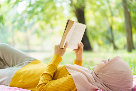 Beautiful Asian muslim woman relaxing in the park under the tree, cheerful muslim woman laying - sitting on the ground and reading a book. Modern lifestyle of muslim people concept.の写真素材