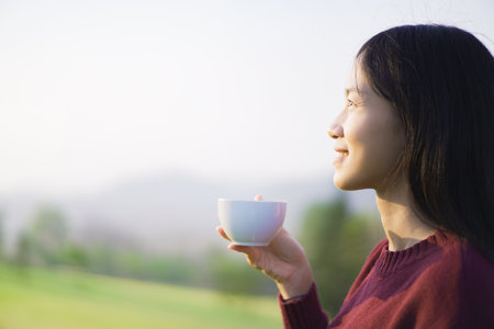 Happy beautiful Asian young woman sitting on the grass field and enjoy relaxing with a stunning view of mountain and forest landscape. Asian young woman drinking a cup of coffee in morning.の写真素材
