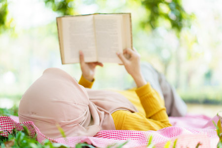 Beautiful Asian muslim woman relaxing in the park under the tree, cheerful muslim woman laying - sitting on the ground and reading a book. Modern lifestyle of muslim people concept.の写真素材