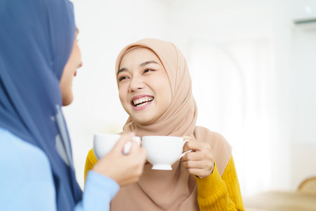 Happy cheerful Asian muslim women drinking a tea or coffee together in the living room and having a business discussion. Modern muslim people lifestyle concept.の写真素材