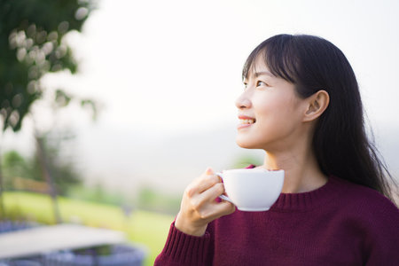 Happy beautiful Asian young woman sitting on the grass field and enjoy relaxing with a stunning view of mountain and forest landscape. Asian young woman drinking a cup of coffee in morning.の写真素材