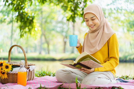 Happy cheerful Asian beautiful muslim woman sitting on the ground at the park and drinking a cup of tea in afternoon. Beautiful muslim woman relaxing and picnic in weekend.の写真素材