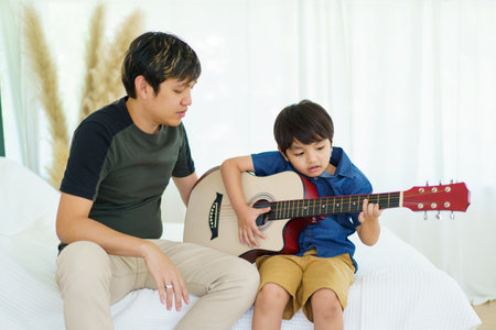 Asian little boy playing an acoustic guitar with his father, a man teaching and playing a guitar with his son in weekend. Happy family - fatherhood concept. Dad teaches his son play a guitar.の写真素材