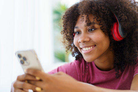 Happy cheerful American - African black teenager woman relaxing in the living room and using smartphone to browse the social network or internet. Woman relaxing in living room and using smartphone.の写真素材