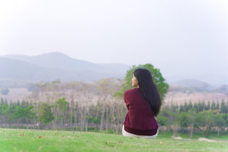 Happy beautiful Asian young woman sitting on the grass field and enjoy relaxing with a stunning view of mountain and forest landscape. Asian young woman drinking a cup of coffee in morning.の写真素材
